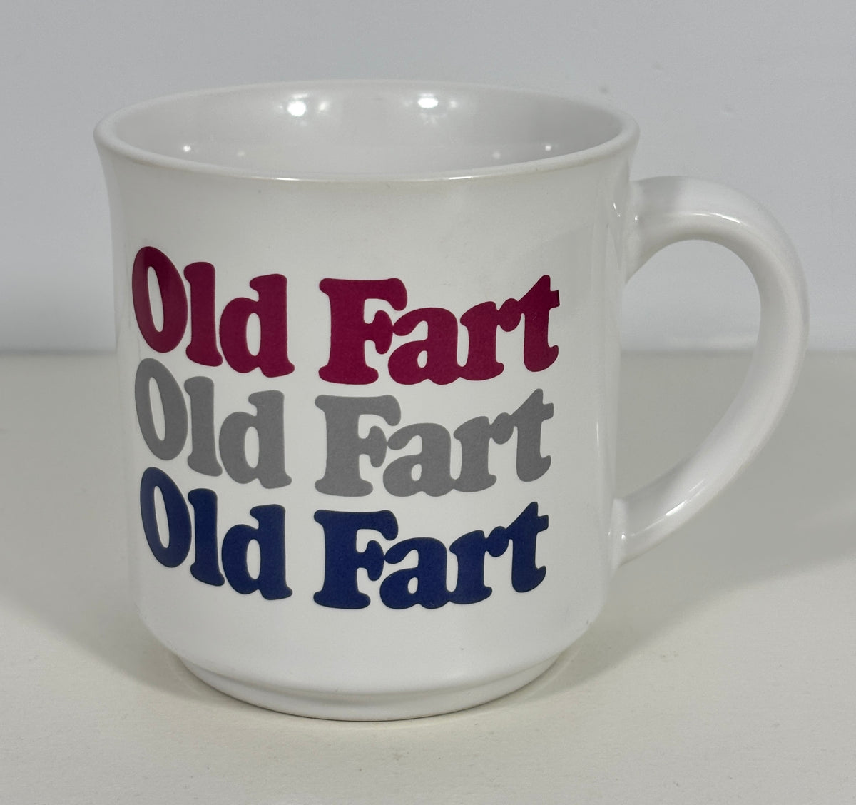 A white ceramic coffee mug with the phrase 'Old Fart' printed three times in alternating dark red, gray, and navy blue fonts, set against a plain light gray background.