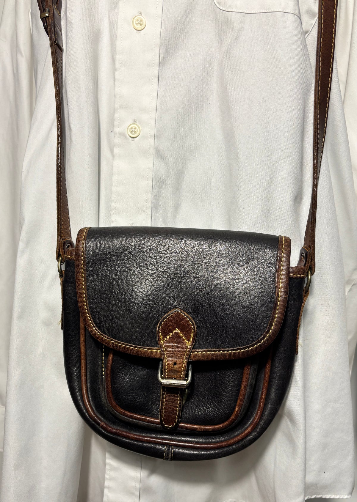 A dark brown leather crossbody bag with brown trim and a silver buckle, worn over a white button-down shirt.