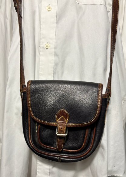 A dark brown leather crossbody bag with brown trim and a silver buckle, worn over a white button-down shirt.