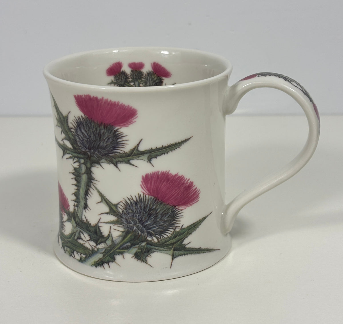 A white ceramic coffee mug decorated with a detailed botanical print of purple thistles and pink flowers, set against a plain light gray background.