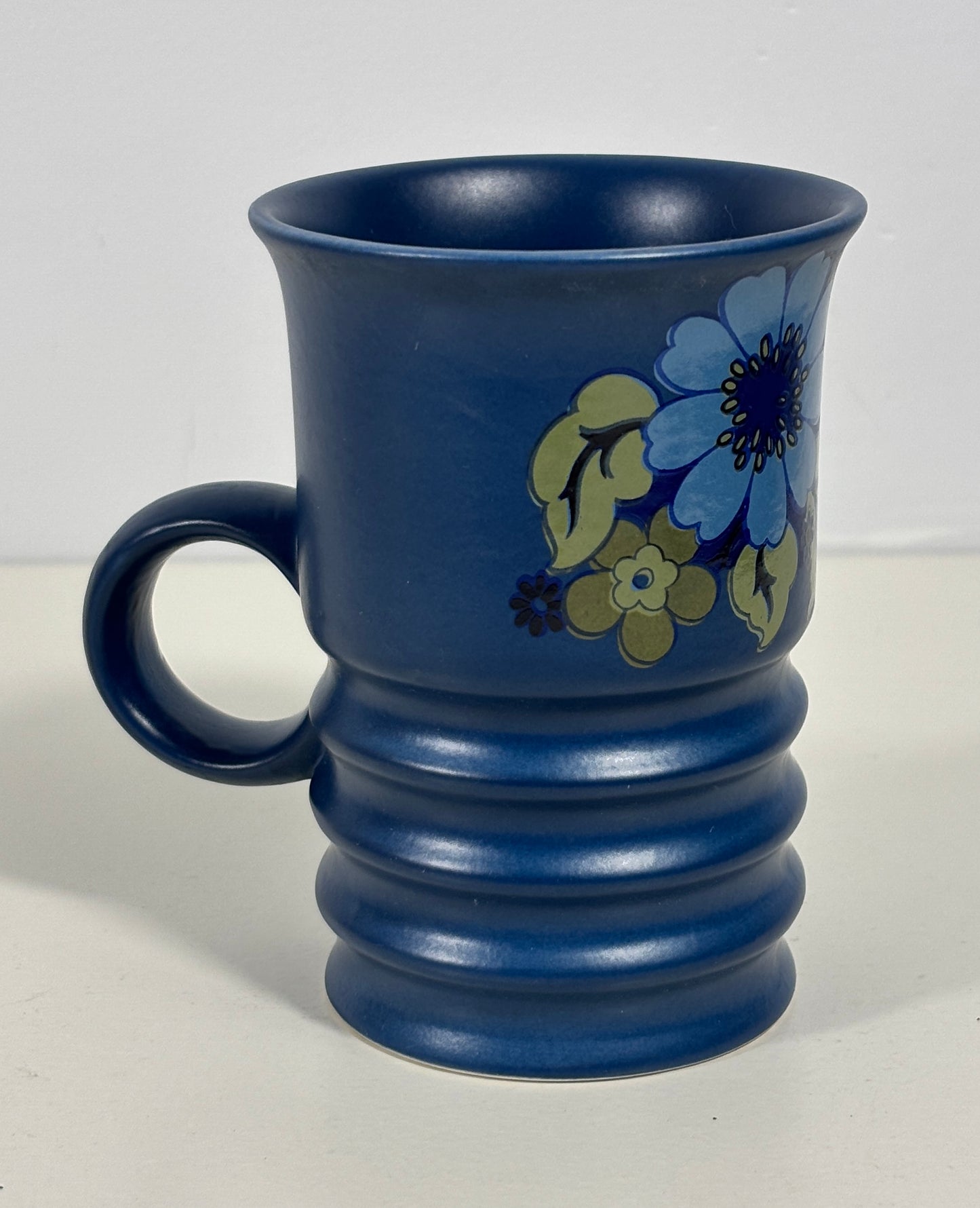 A cobalt blue ceramic mug with a ribbed base and a floral pattern featuring blue, yellow, and green flowers, set against a plain light background.