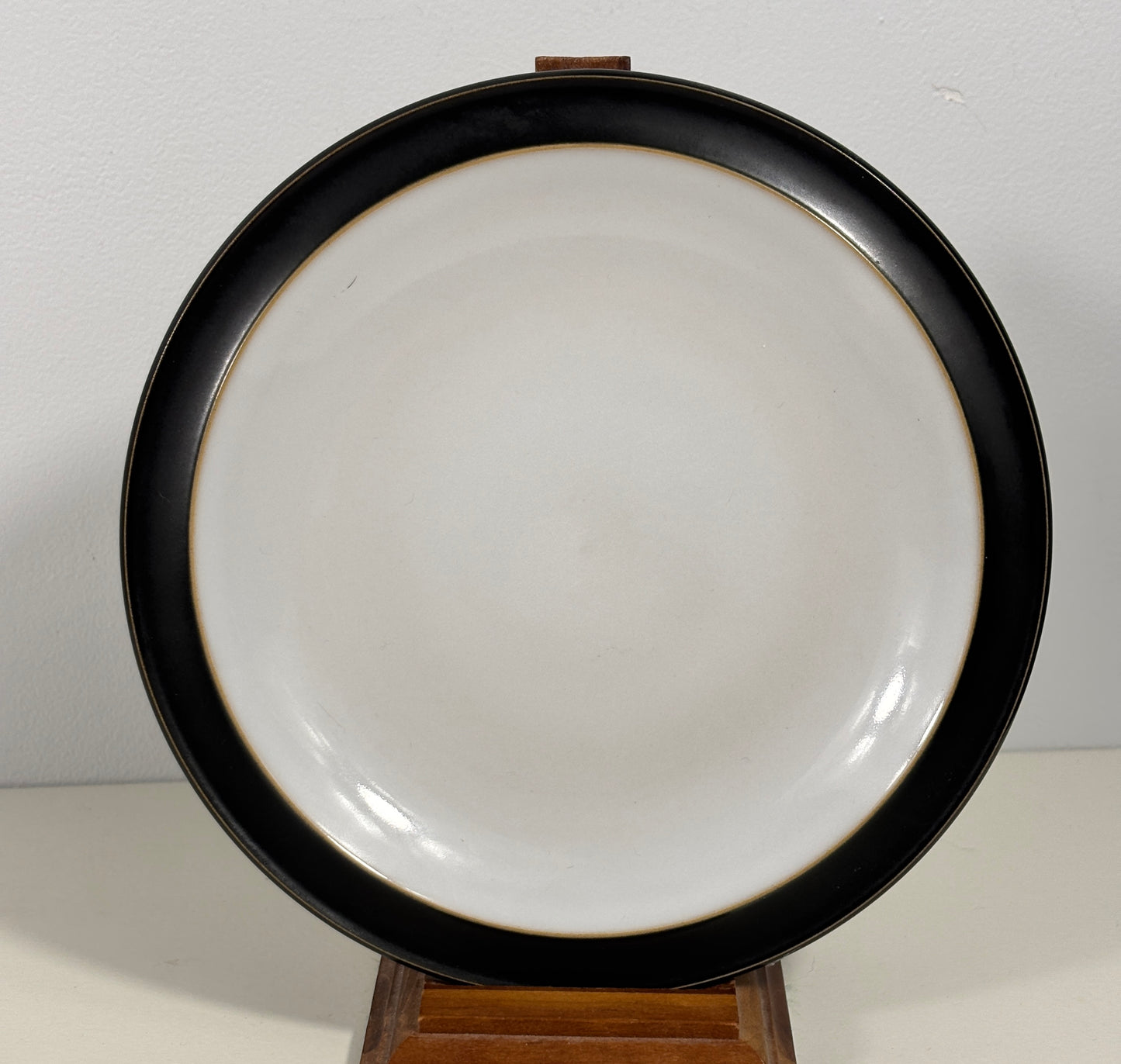 A round ceramic plate with a wide black rim and a glossy white center, displayed on a small wooden stand against a plain light background.