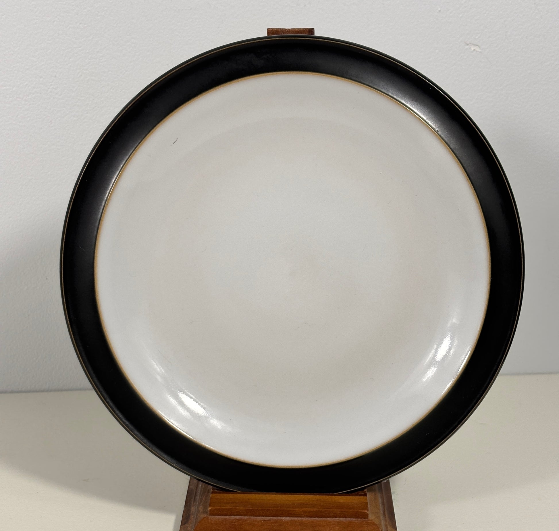 A round ceramic plate with a wide black rim and a glossy white center, displayed on a small wooden stand against a plain light background.