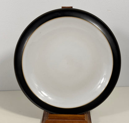 A round ceramic plate with a wide black rim and a glossy white center, displayed on a small wooden stand against a plain light background.