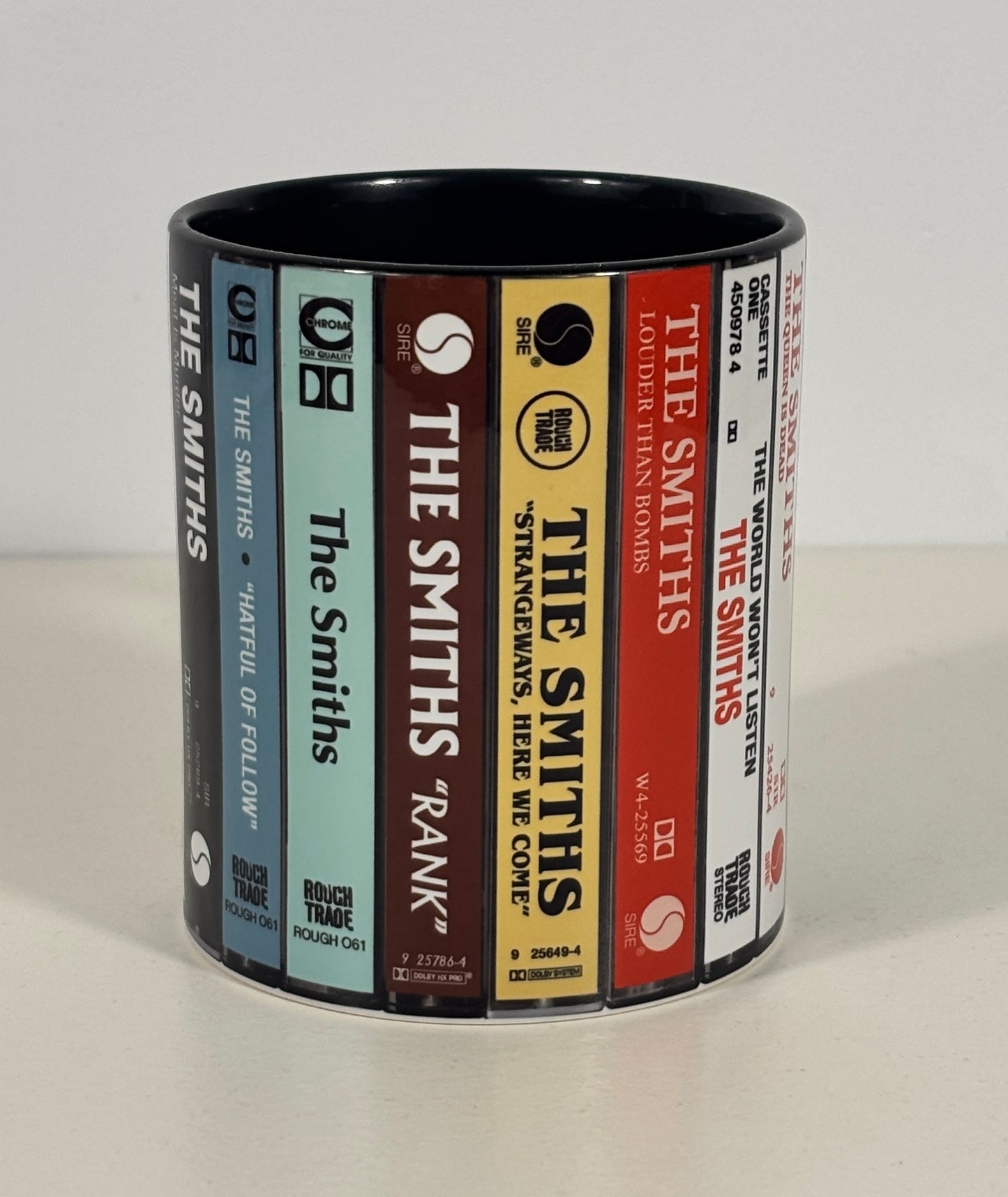 A cylindrical ceramic mug printed with a wraparound design resembling book spines, including titles such as The Smiths, Stranger Things, and The Godfather. The mug is photographed on a plain light background.