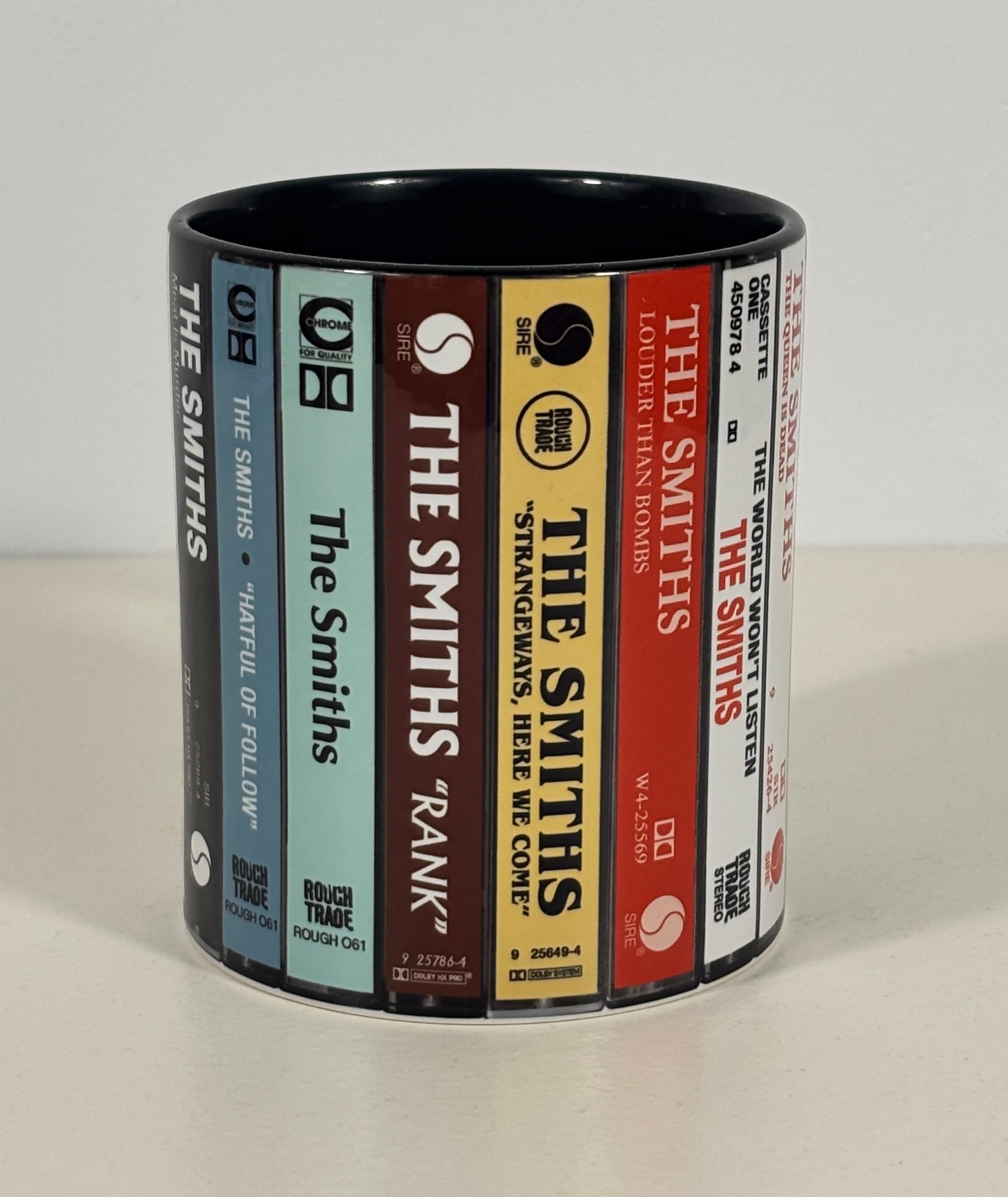 A cylindrical ceramic mug printed with a wraparound design resembling book spines, including titles such as The Smiths, Stranger Things, and The Godfather. The mug is photographed on a plain light background.