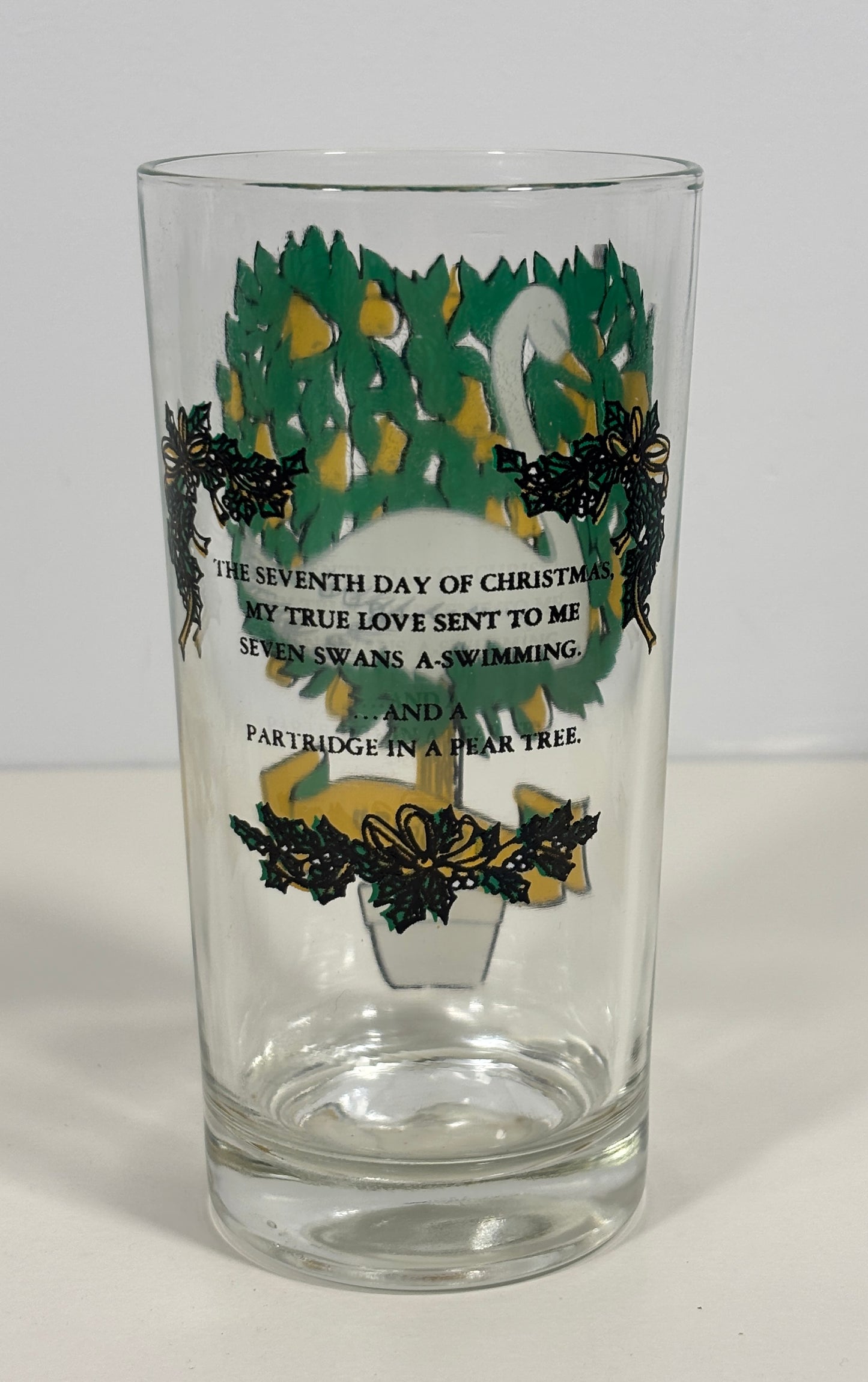 A clear glass drinking tumbler featuring a Christmas tree design with green and yellow ornaments, black holly leaves, and black printed text about the seventh day of Christmas and seven swans swimming in a pear tree.