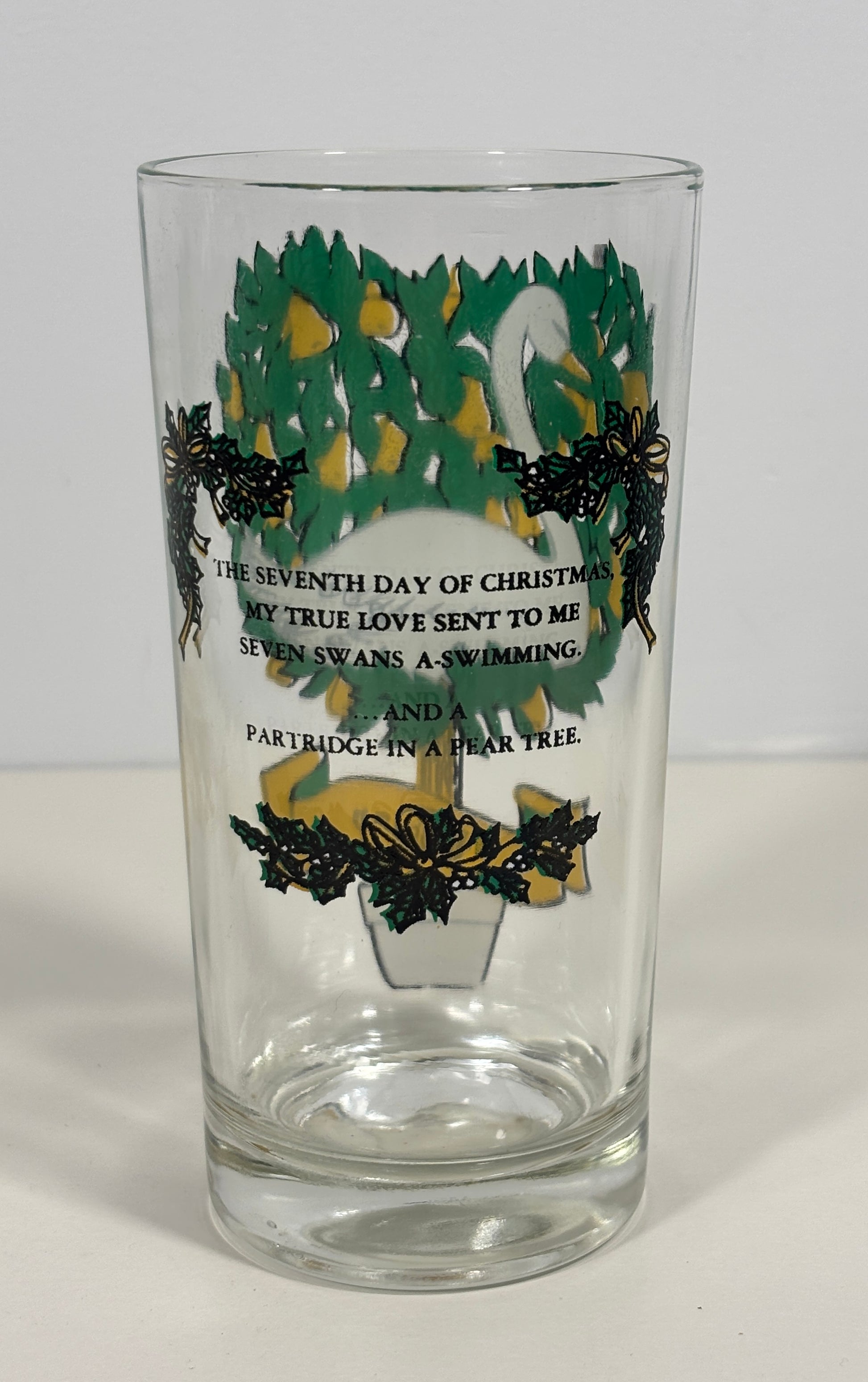 A clear glass drinking tumbler featuring a Christmas tree design with green and yellow ornaments, black holly leaves, and black printed text about the seventh day of Christmas and seven swans swimming in a pear tree.