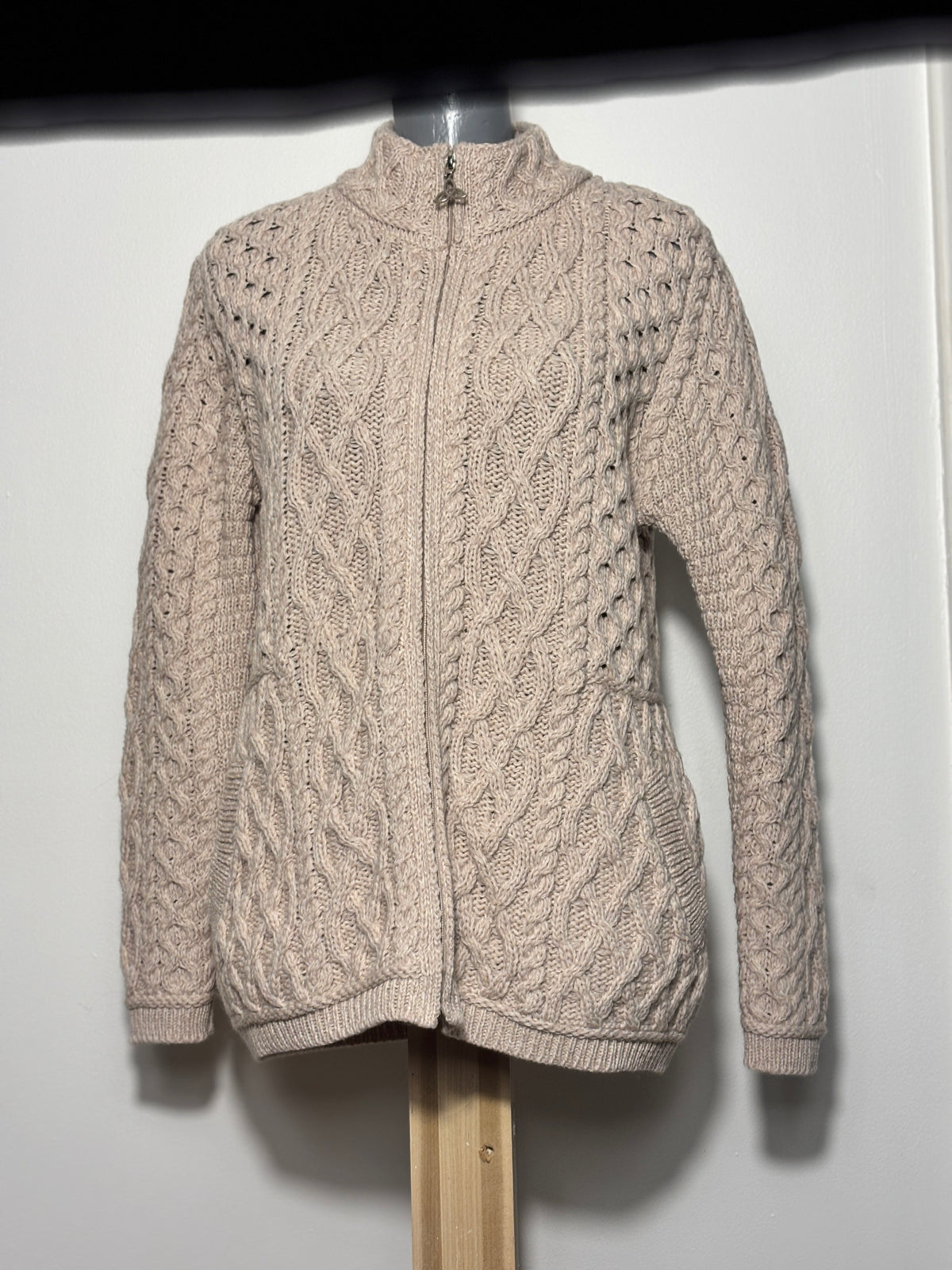 A cable-knit zip-up sweater in a light oatmeal beige color, displayed on a mannequin torso against a plain white wall.