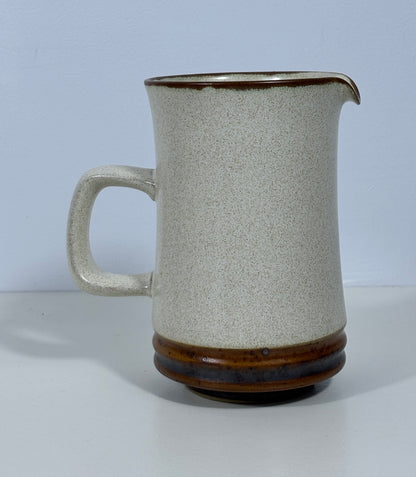 A handmade ceramic pitcher with a speckled off-white body, a dark brown glazed rim, and a matching dark brown glazed base, set against a plain light gray background.