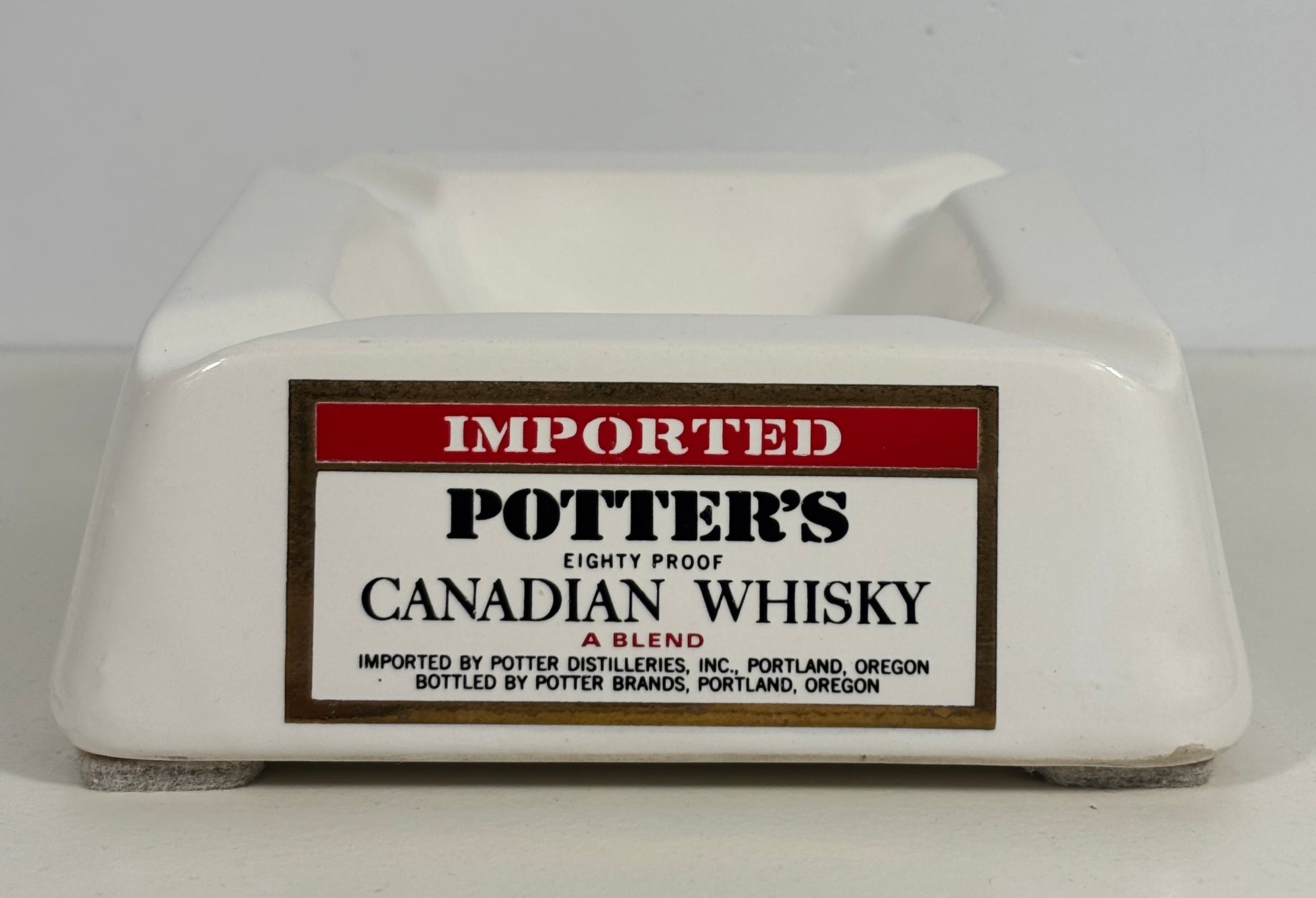 A white ceramic potter's ashtray featuring a vintage-style label that reads 'IMPORTED POTTER'S CANADIAN WHISKY'.