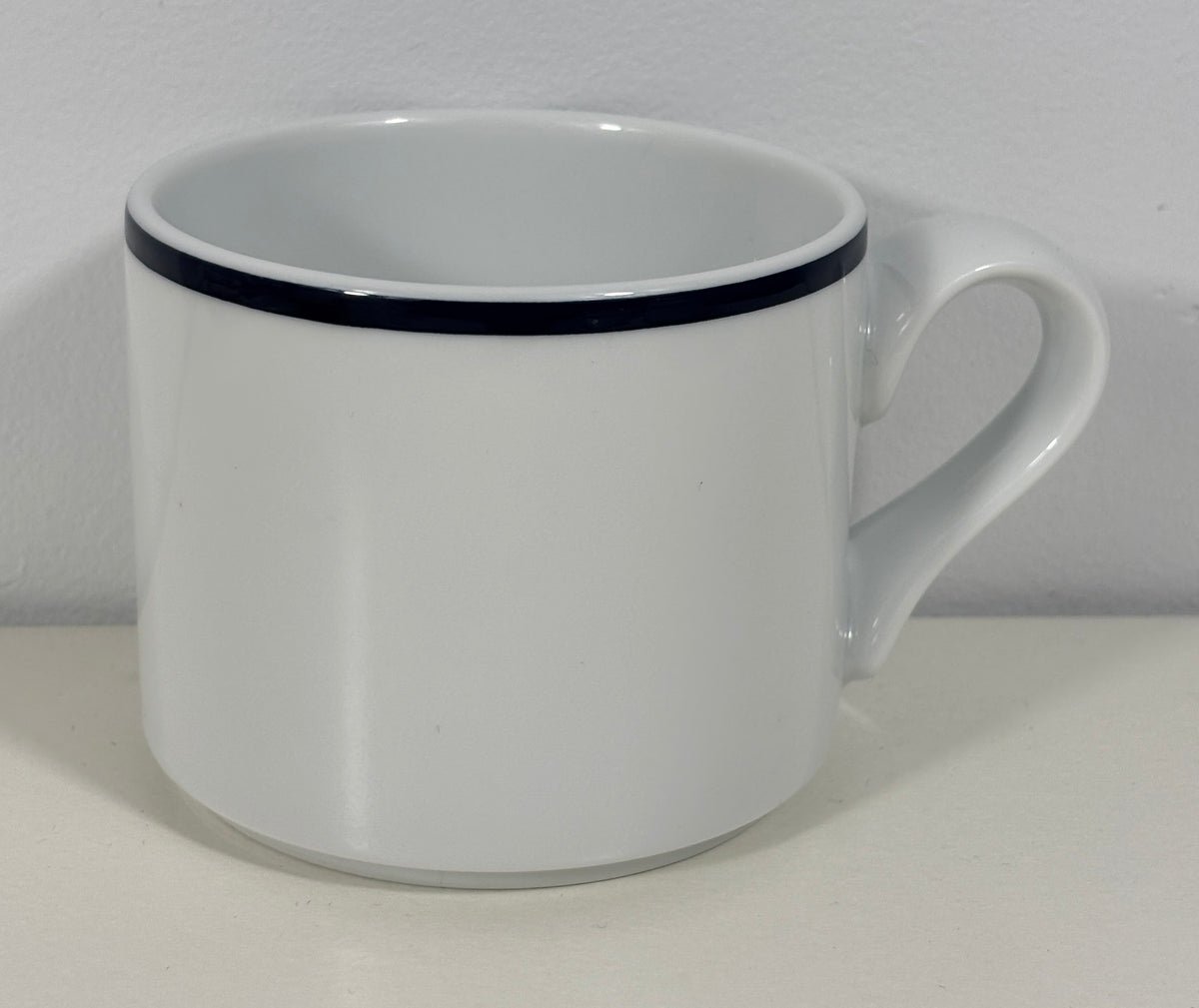 A white ceramic coffee mug with a single thin black stripe around the rim, shown on a plain white surface against a white wall.