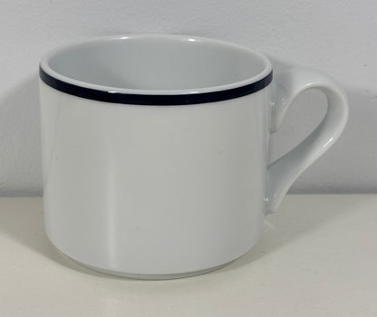 A white ceramic coffee mug with a single thin black stripe around the rim, shown on a plain white surface against a white wall.