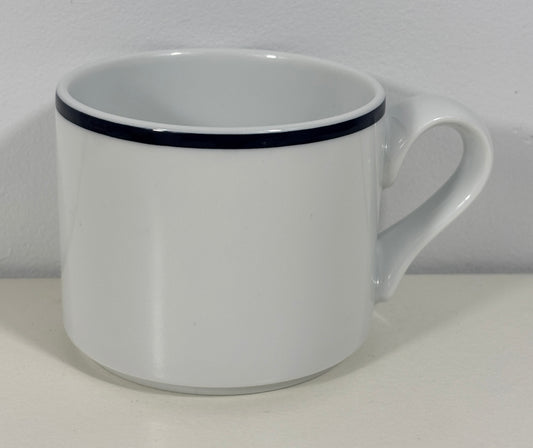 A white ceramic coffee mug with a single thin black stripe around the rim, shown on a plain white surface against a white wall.