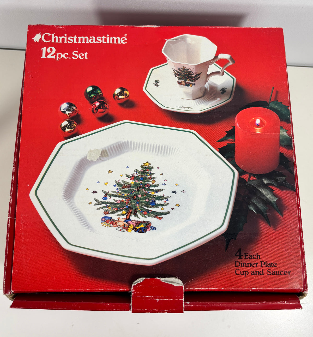 A Christmas-themed 12-piece dinnerware set in its original red packaging box. The box features illustrations of a white octagonal plate and matching cup and saucer decorated with a green Christmas tree, alongside red ornaments, a red candle, and green leaves.
