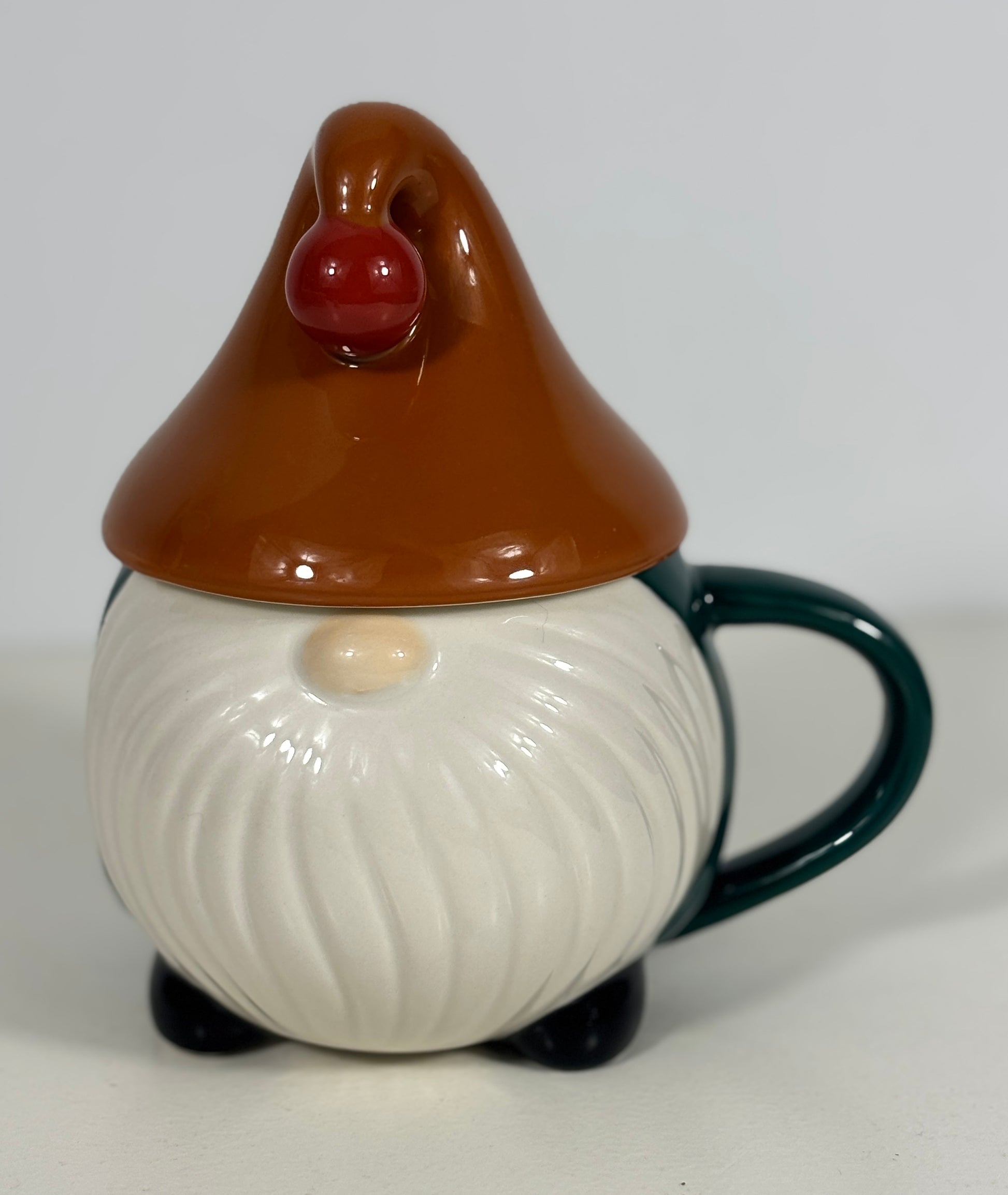 A ceramic gnome mug with a matching lid. The gnome has a brown pointed hat with a red nose, a white beard, and a dark green handle and feet, set against a plain light gray background.