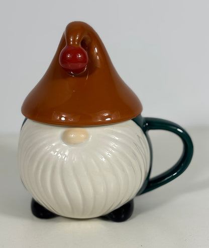 A ceramic gnome mug with a matching lid. The gnome has a brown pointed hat with a red nose, a white beard, and a dark green handle and feet, set against a plain light gray background.