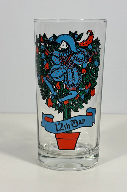 A clear glass pint glass featuring a colorful illustration of a jester in a blue suit surrounded by green foliage and red berries, with a blue banner reading '12th Bar' at the bottom.