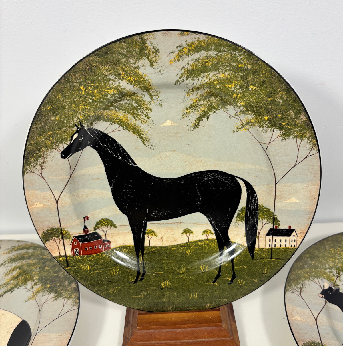 A decorative ceramic plate featuring a black horse in a rural landscape with trees, a red barn, and a small house, displayed on a wooden stand against a white wall.