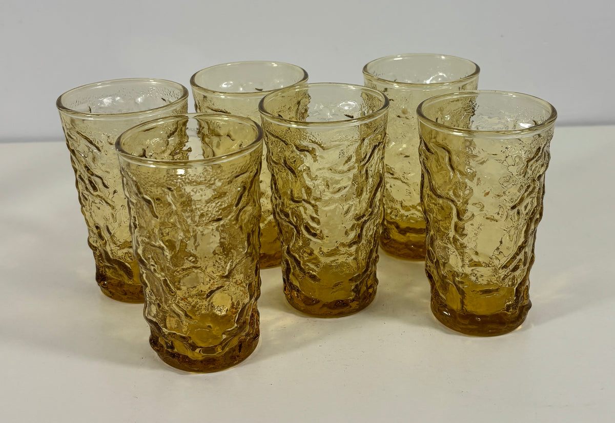 Six amber-tinted textured glass tumblers arranged on a white surface against a light gray background.