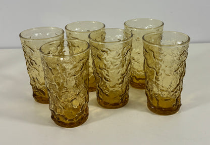 Six amber-tinted textured glass tumblers arranged on a white surface against a light gray background.