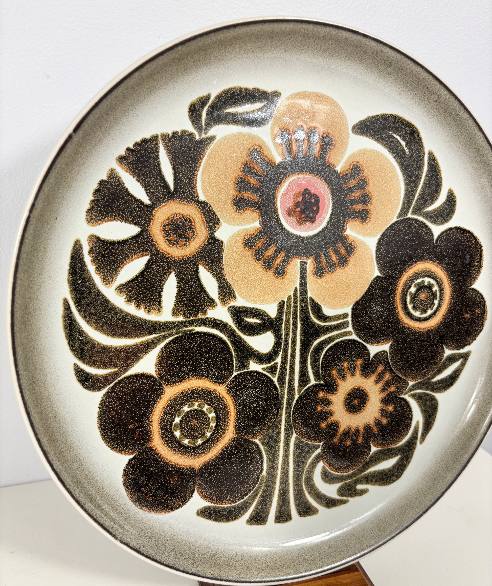 A round ceramic plate featuring a retro floral pattern with dark brown flowers and leaves on a cream background, with a small pink accent in the center flower.