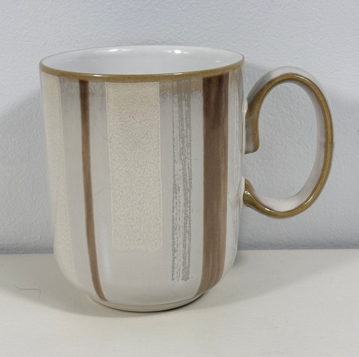 A ceramic mug with a white and beige textured glaze, featuring vertical brown and gray brushstroke patterns and a gold-toned rim and handle.