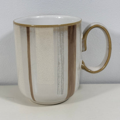 A ceramic mug with a white and beige textured glaze, featuring vertical brown and gray brushstroke patterns and a gold-toned rim and handle.