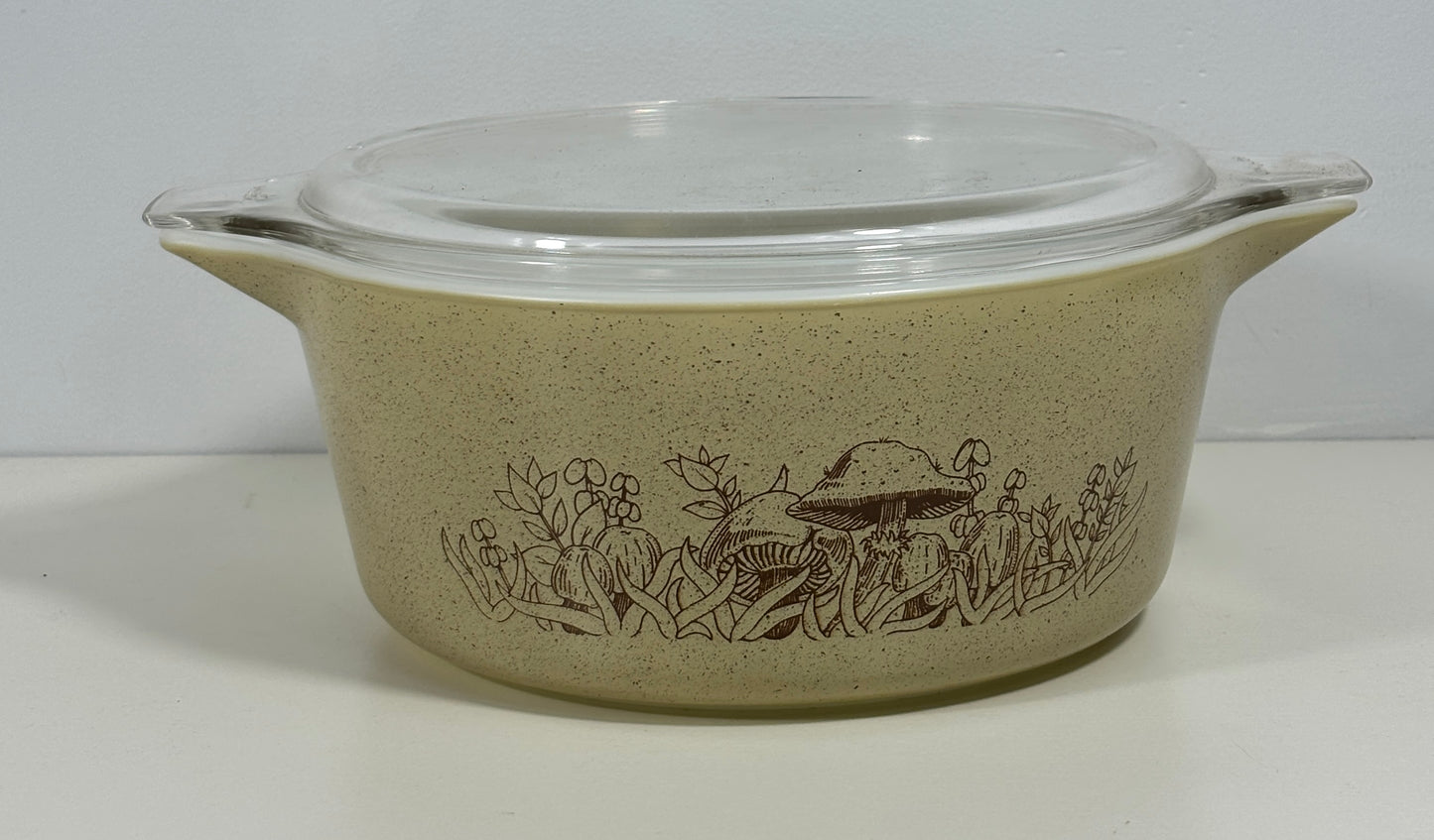 A vintage oval ceramic casserole dish with a clear glass lid, featuring a brown mushroom and floral border pattern on a speckled beige background.