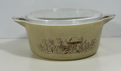 A vintage oval ceramic casserole dish with a clear glass lid, featuring a brown mushroom and floral border pattern on a speckled beige background.