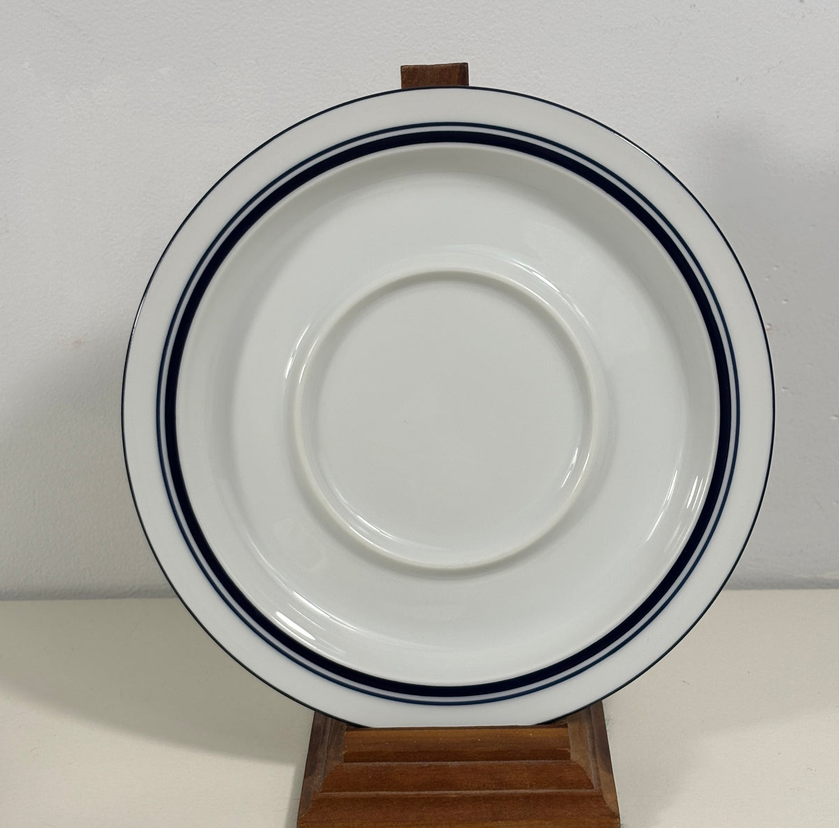 A white ceramic plate with two dark blue stripes near the rim, displayed on a small wooden stand against a plain light background.