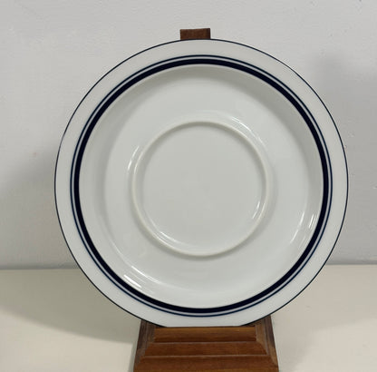 A white ceramic plate with two dark blue stripes near the rim, displayed on a small wooden stand against a plain light background.