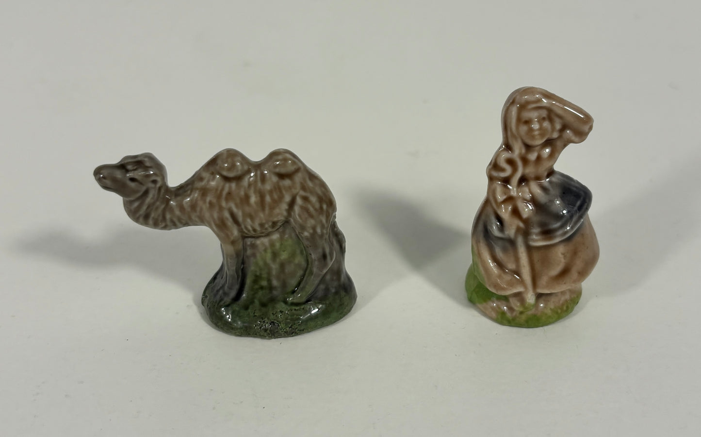 Two small glazed ceramic figurines on a plain light background: a camel standing on the left and a seated robed figure on the right.
