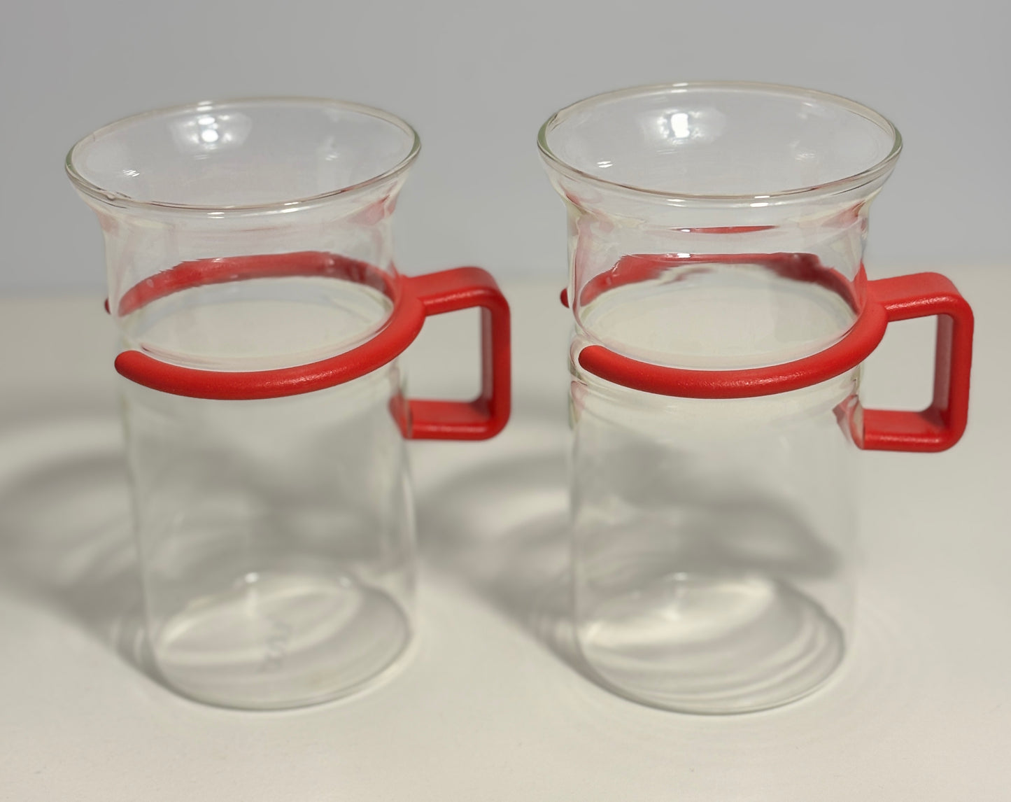 Two clear glass beakers with red plastic handles, shown side by side on a light gray background.