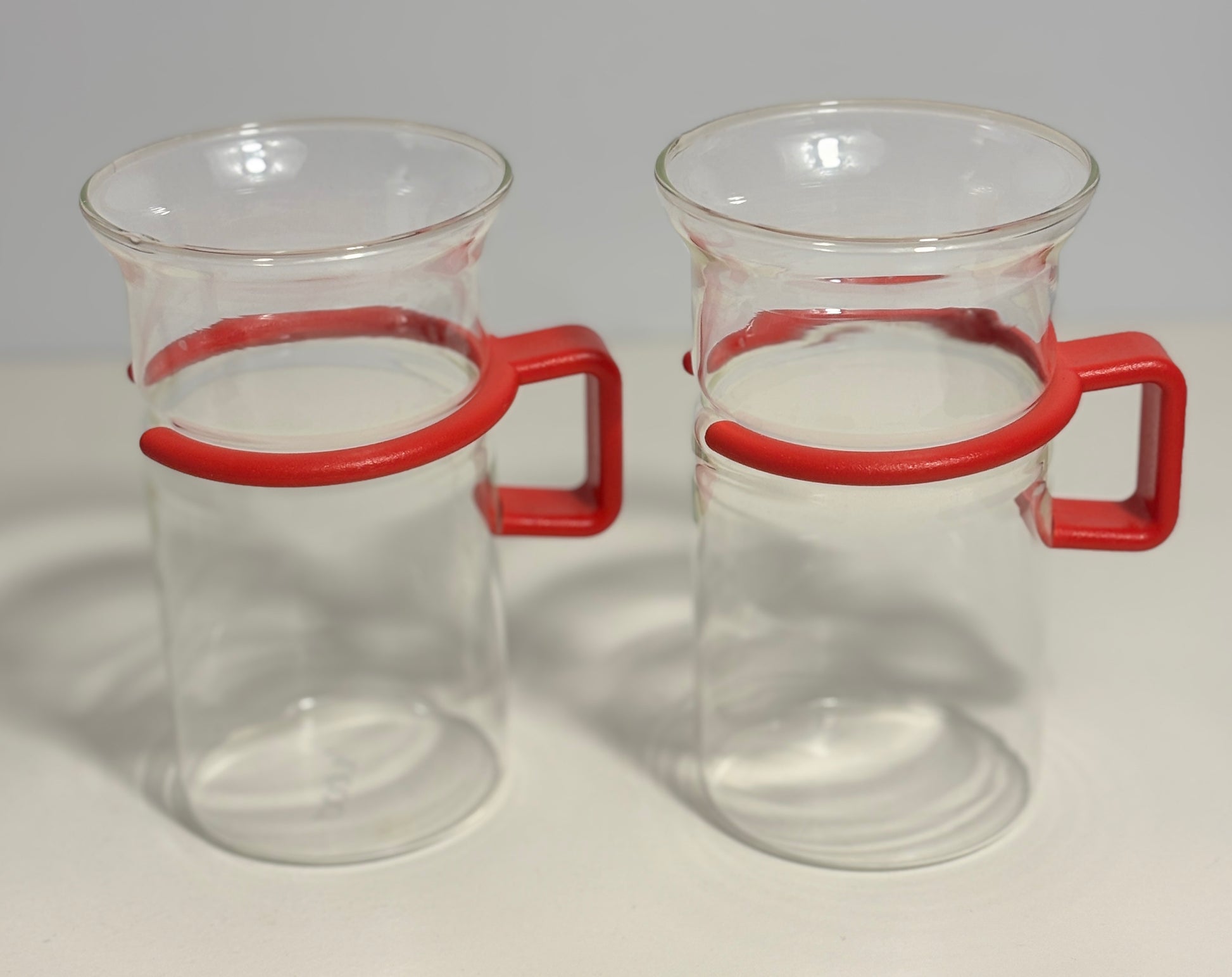 Two clear glass beakers with red plastic handles, shown side by side on a light gray background.