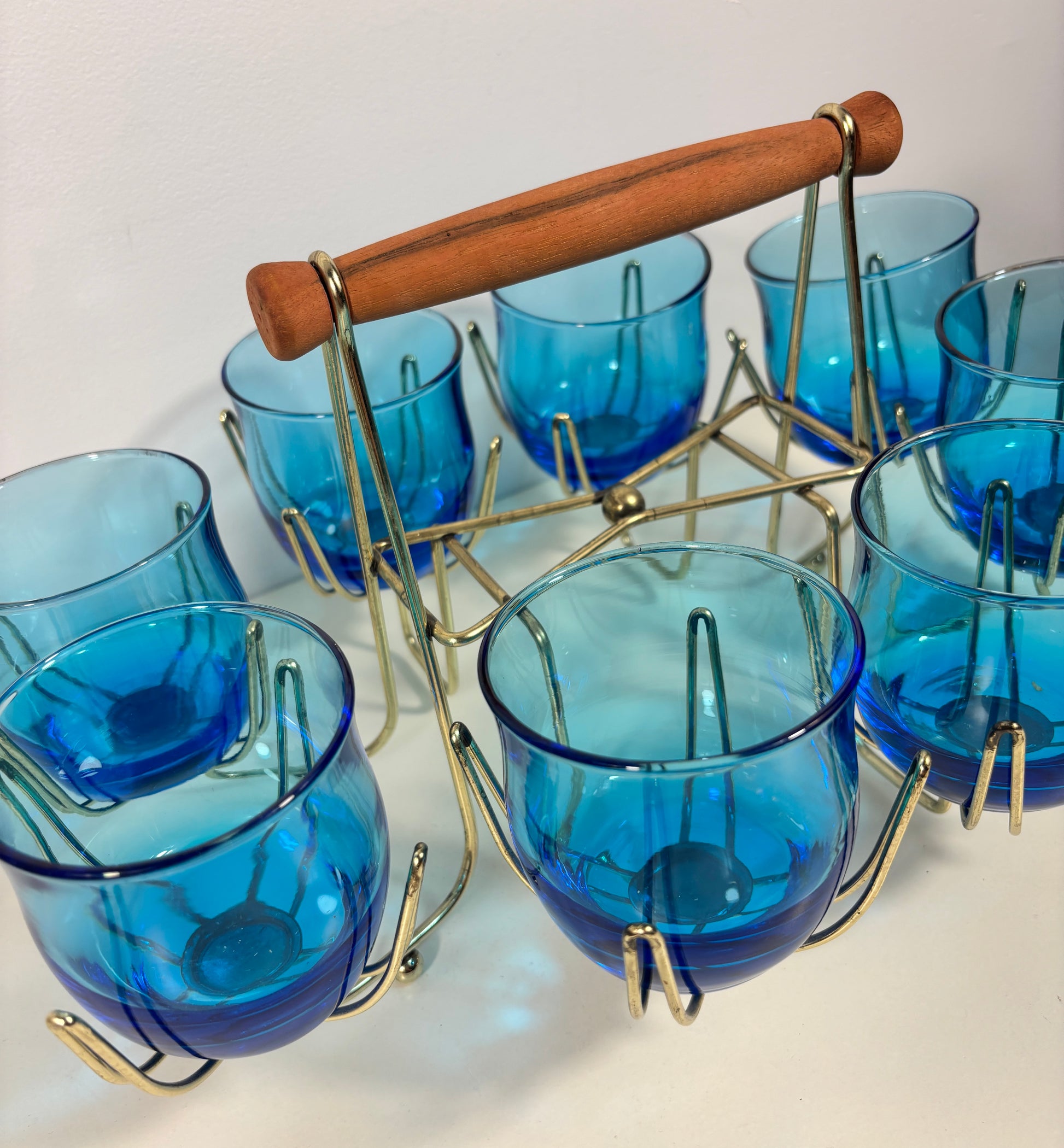 A set of six cobalt blue glass teacups arranged in a brass cup carrier with a wooden handle, photographed on a plain light background.