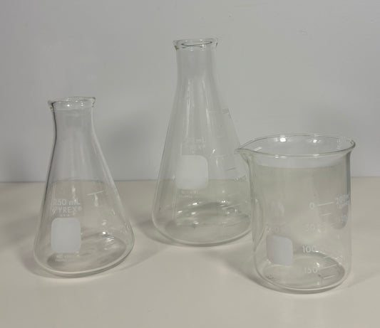 Three clear glass laboratory glassware pieces on a white surface against a light gray background: an Erlenmeyer flask, a conical flask, and a beaker, each with a frosted square label.