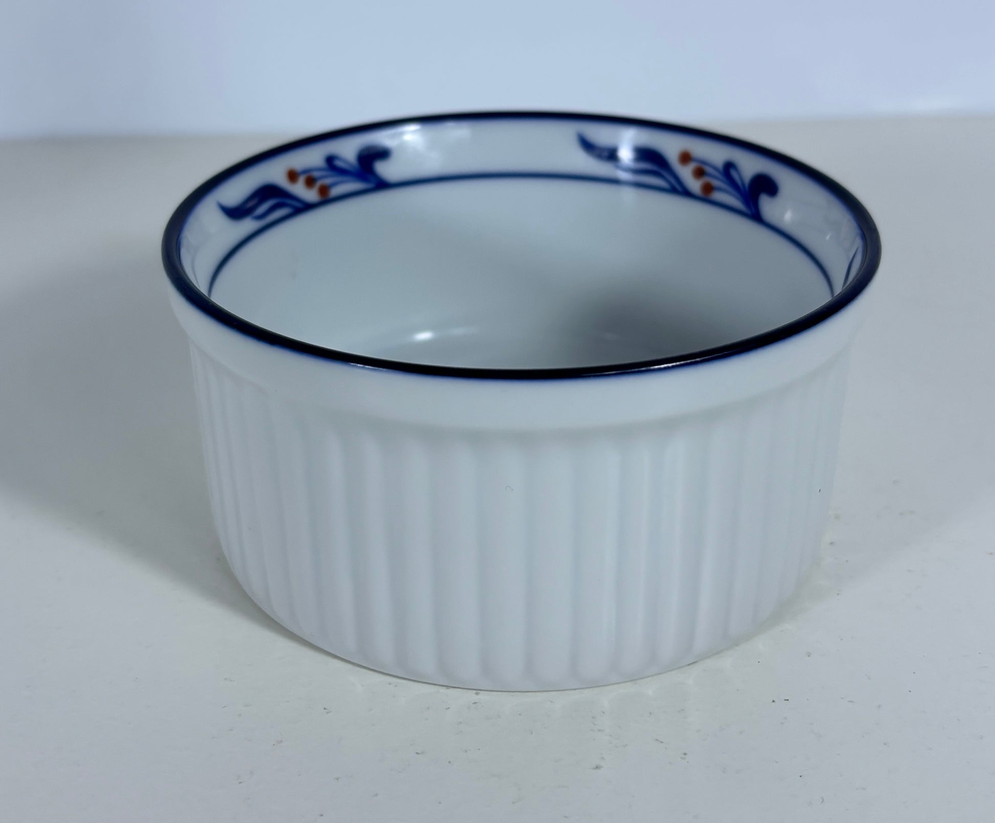 A white ribbed ceramic ramekin with a blue rim and a delicate blue and orange floral pattern around the inner edge, shown on a light gray surface.