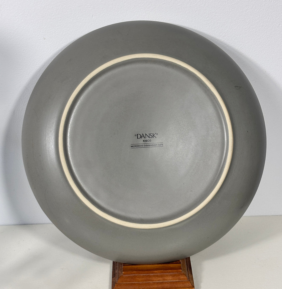A round gray ceramic plate with a cream-colored inner rim, displayed on a wooden stand against a plain white background. The brand name 'DANSK' is printed in the center.