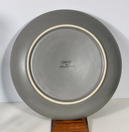 A round gray ceramic plate with a cream-colored inner rim, displayed on a wooden stand against a plain white background. The brand name 'DANSK' is printed in the center.