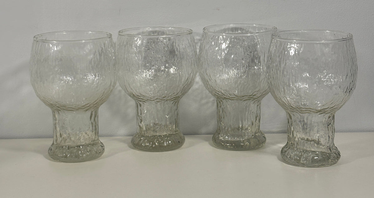 Four clear textured glass tumblers with flared bases, arranged side by side on a white surface against a white background.