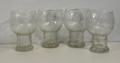 Four clear textured glass tumblers with flared bases, arranged side by side on a white surface against a white background.