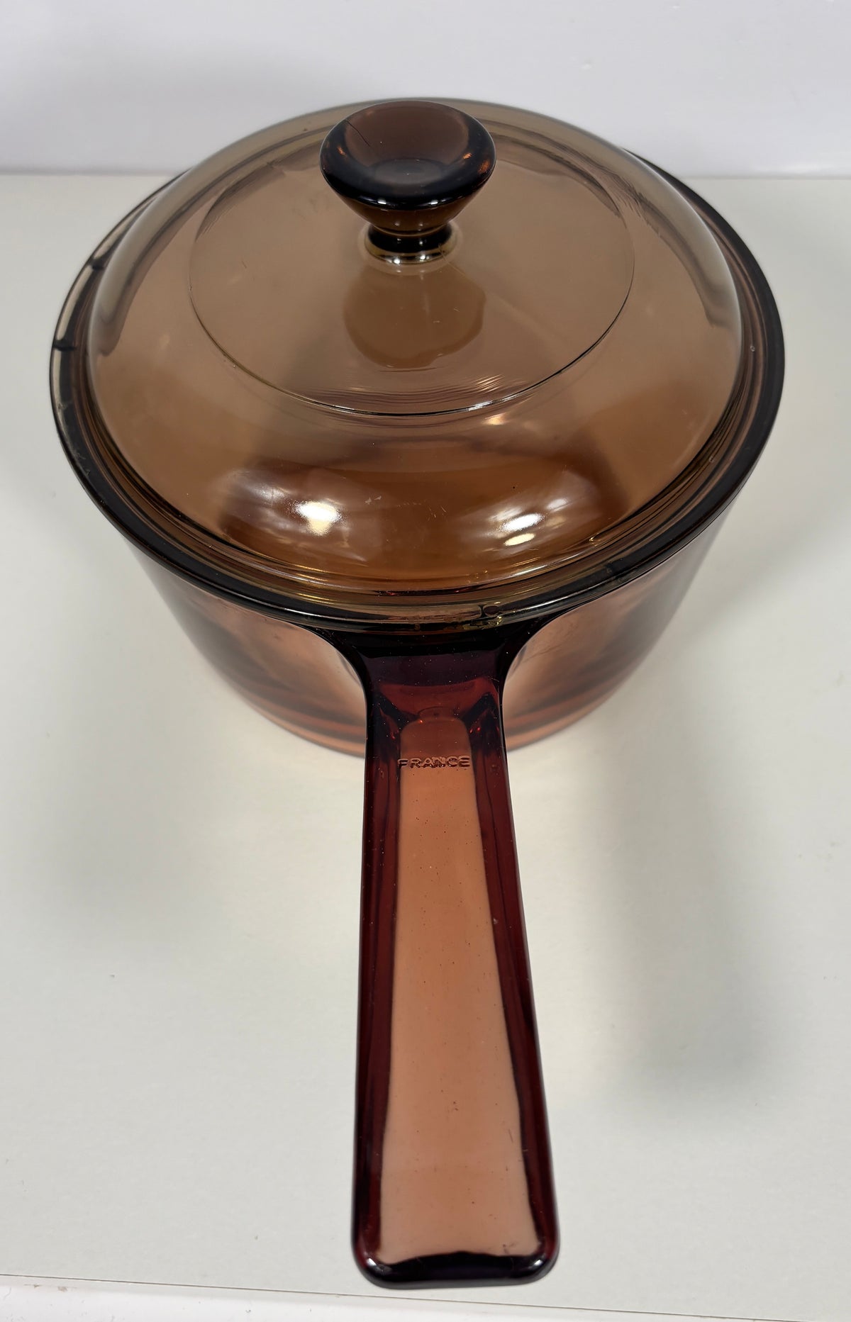 A translucent amber-brown glass saucepan with a matching lid and long handle, photographed on a plain light background.