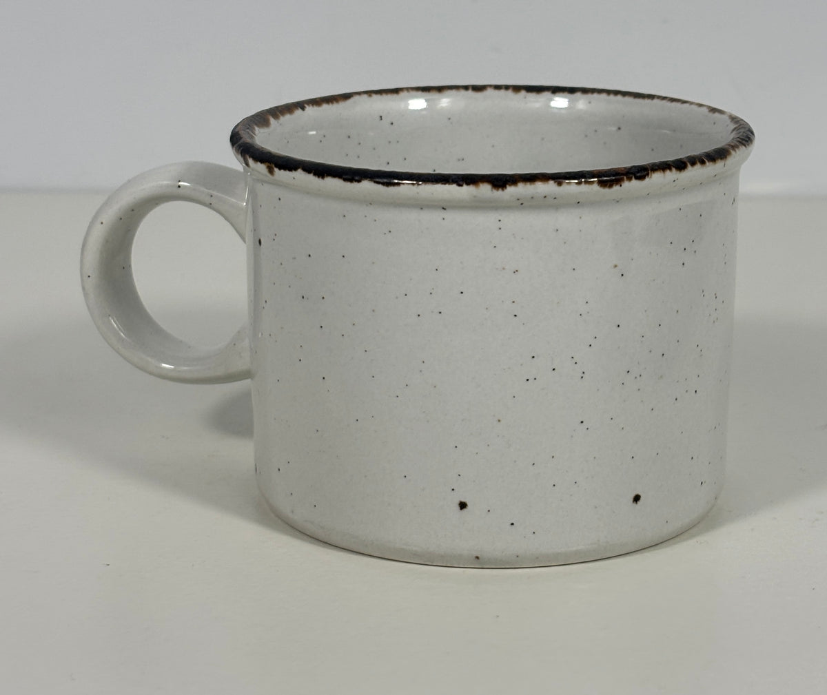 A white speckled ceramic mug with a dark brown rim, shown on a plain light gray background.