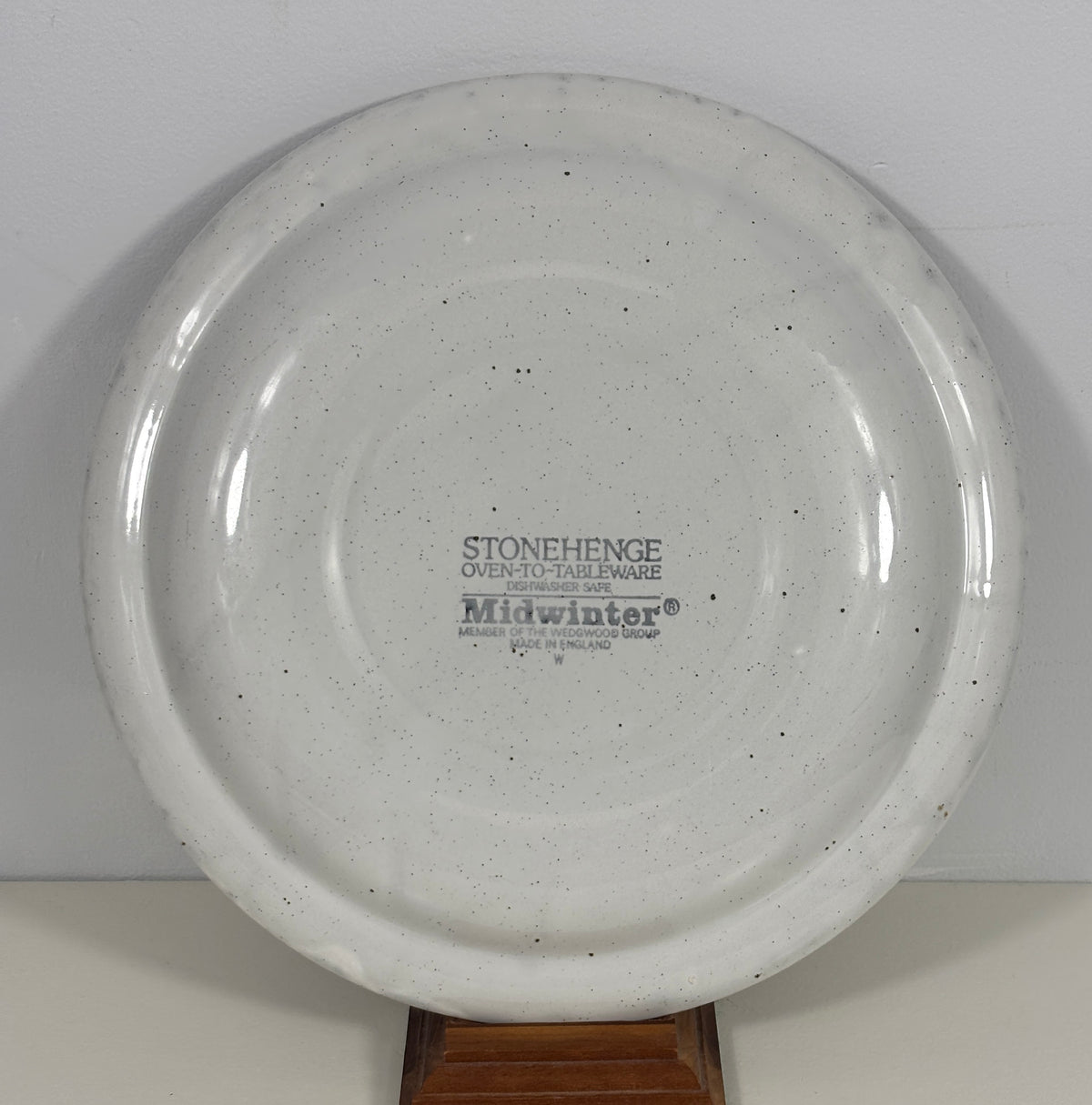 A round off-white speckled ceramic plate or shallow dish displayed on a wooden stand against a plain light background. The center is printed with the brand name 'Midwinter' and product text.