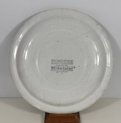 A round off-white speckled ceramic plate or shallow dish displayed on a wooden stand against a plain light background. The center is printed with the brand name 'Midwinter' and product text.