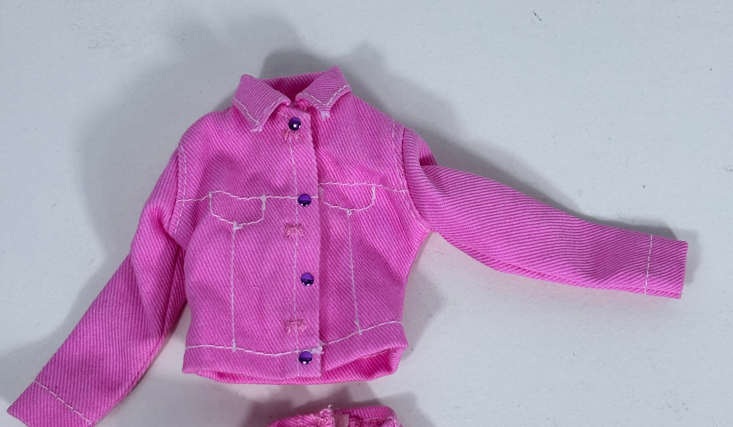 A pink doll jacket with white contrast stitching and purple buttons, laid flat on a light gray background.