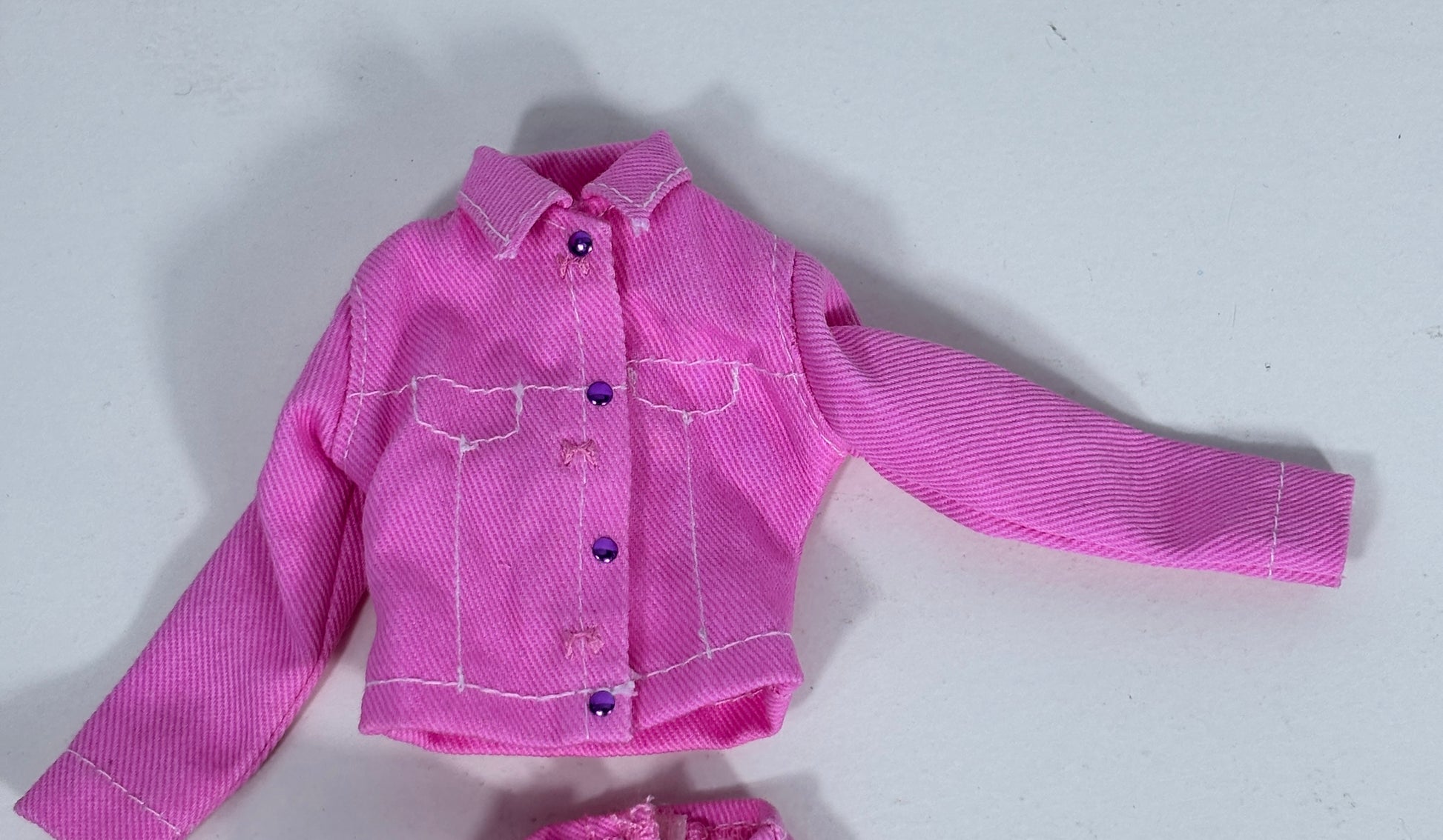 A pink doll jacket with white contrast stitching and purple buttons, laid flat on a light gray background.