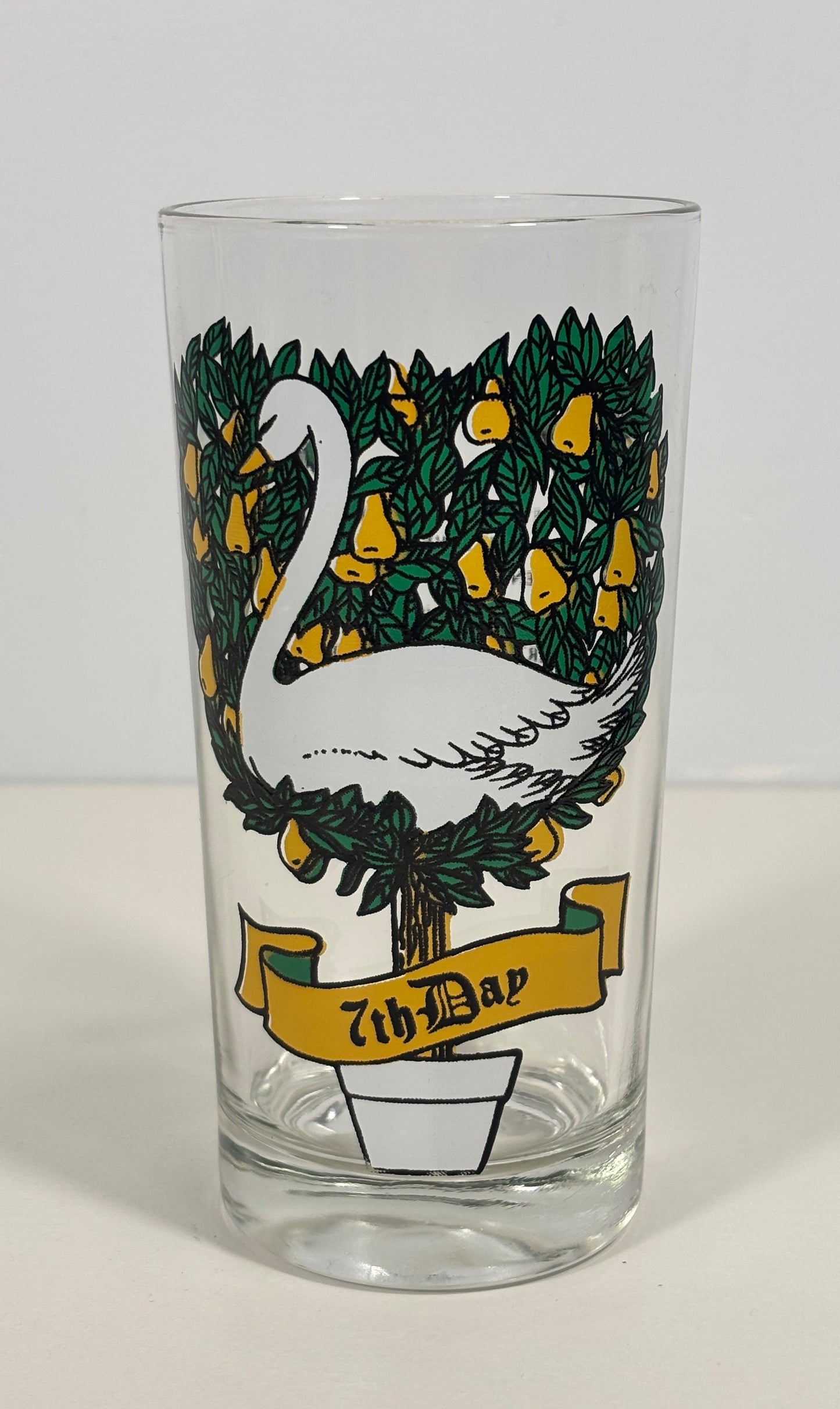 A clear glass drinking tumbler featuring a printed design of a white swan on a tree with green leaves and yellow fruit, with a yellow banner reading '7th Day' near the bottom, shown against a plain light background.