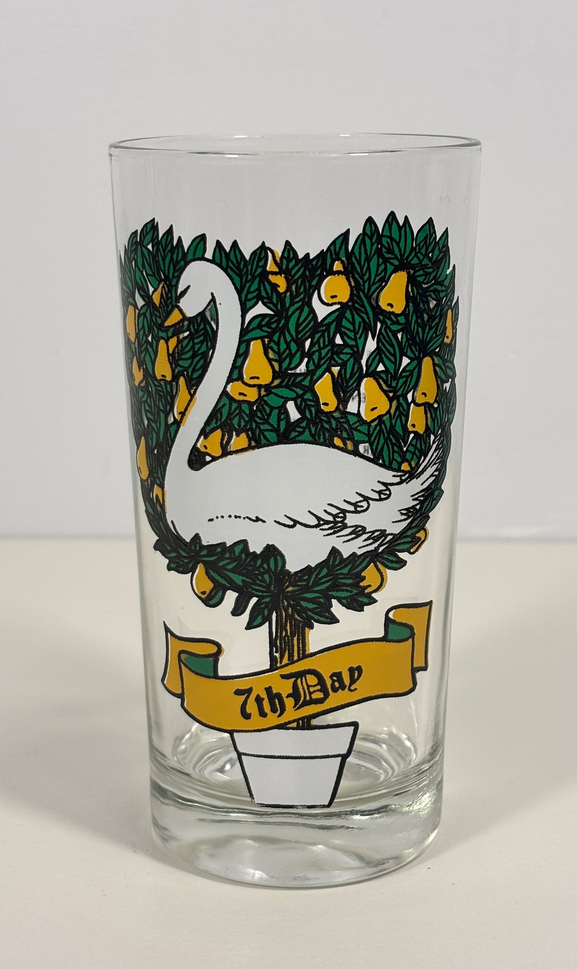 A clear glass drinking tumbler featuring a printed design of a white swan on a tree with green leaves and yellow fruit, with a yellow banner reading '7th Day' near the bottom, shown against a plain light background.